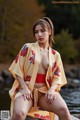 A woman in a yellow kimono sitting on a rock by the water.