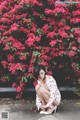 A woman sitting on the ground in front of a bush of flowers.